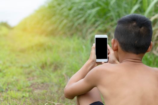 The boy is sitting on the grass and playing mobile phone. Background of grass and sunlight.