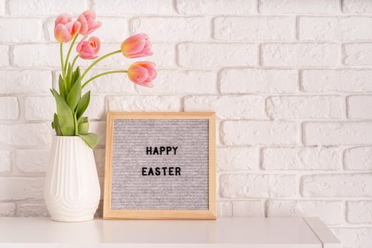 Easter hliday concept. Vase with tulips and gray felt letter board with words Happy Easter on white bricks background