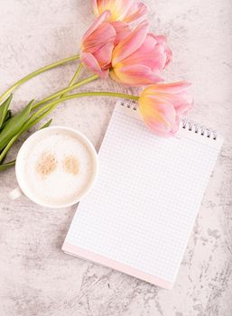 Cup of cappuccino, notebook and pink tulips top view on marble background