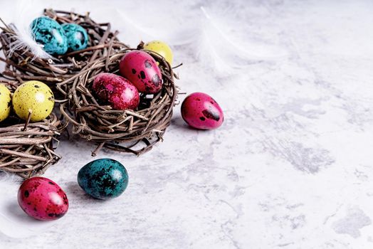 Easter holiday concept. Colored Easter quail eggs with feathers in a nest on gray marble background with copy space