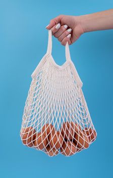 Easter concept. No plastic bag concept. Minimal concept. Female hand holding white mesh shopping bag with brown chicken eggs on blue background top view flat lay with copy space
