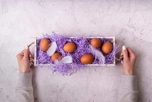 Easter holiday concept. Woman hands holding white wooden box with chicken eggs on white marble background top view flat lay