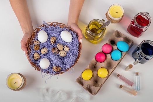 Easter holiday concept. Easter craft. Woman hands holding a big basket with quail and chicken eggs decorated with feathers and colorful water top view flat lay