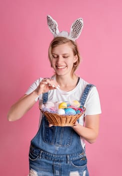 Easter holiday concept. Young woman in bunny ears putting a feather into the basket with colored Easter eggs isolated on pink background