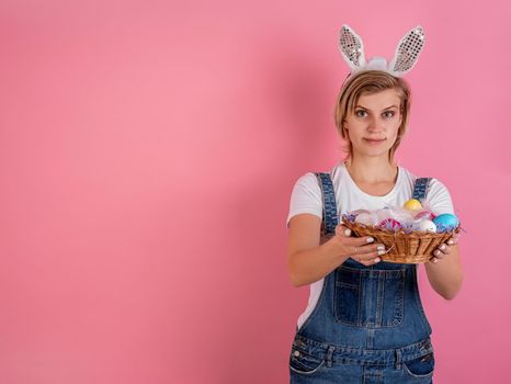 Easter holiday concept. Young woman in bunny ears holding basket with colored Easter eggs isolated on pink background with copy space