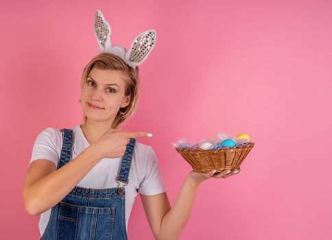 Easter holiday concept. Young blond woman in bunny ears pointing to the basket with colored eggs and winking isolated on pink background with copy space