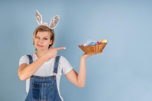 Easter holiday concept. Young blond woman with bunny ears pointing to the basket with colored eggs and winking isolated on blue background with copy space