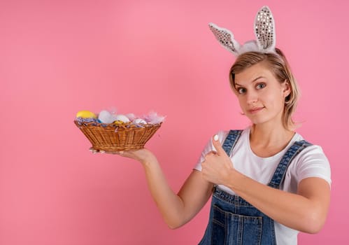 Easter holiday concept. Pretty young woman in bunny ears showing thumbs ups and holding a basket with Easter colored eggs isolated on pink background