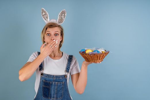 Easter holiday concept. Surprised woman with her hand next to the mouth wearing bunny ears holding a basket with colored eggs isolated on blue background with copy space