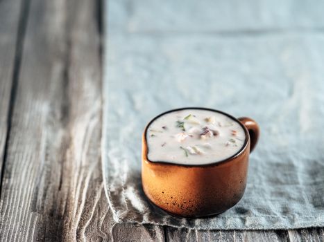 Authentic pakistani cucumber raita sauce on blue linen napkin over gray wooden background. Raita sauce dip with cucumber, onion and tomato add-ins in brown ceramic sauceboat. Copy space