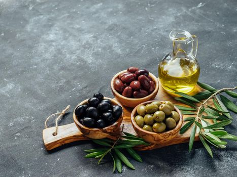 Set of green, black and red or pink olives and olive oil on gray background. Different types of olives in olive wooden bowls and olive oil over wooden cutting board. Copy space