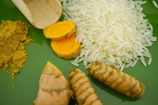 Fresh raw turmeric roots and slices next to heaps of rice and spice powder on a green plate. Selective focus and free space for text