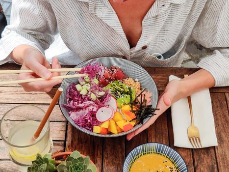 Woman eating tasty colorful healthy natural organic vegetarian Hawaiian poke bowl using asian chopsticks on rustic wooden table. Healthy natural organic eating concept.