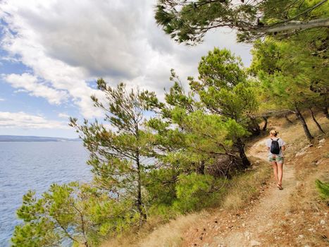 Young active feamle tourist wearing small backpack walking on coastal path among pine trees looking for remote cove to swim alone in peace on seaside in Croatia. Travel and adventure concept.