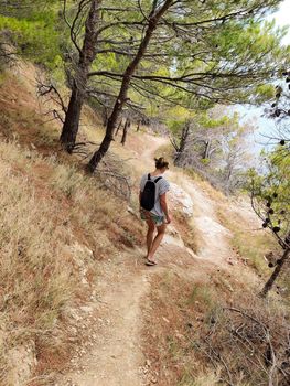 Young active feamle tourist wearing small backpack walking on coastal path among pine trees looking for remote cove to swim alone in peace on seaside in Croatia. Travel and adventure concept.