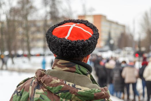 man in camouflage jacket and cossack hat with white cross on red watching blurry crowd of people - view from back.