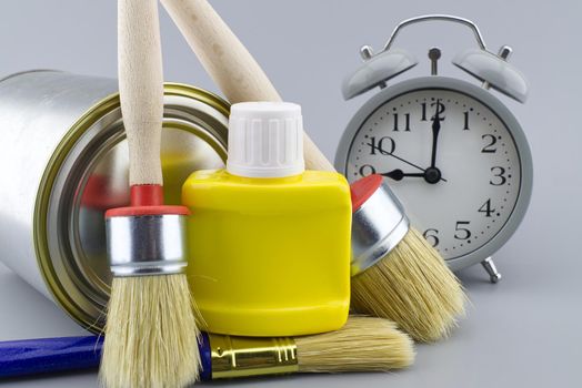 New color, decorating or renovations concept with a still life of an alarm clock with assorted clean paint brushes and unlabeled tin of paint over a grey background