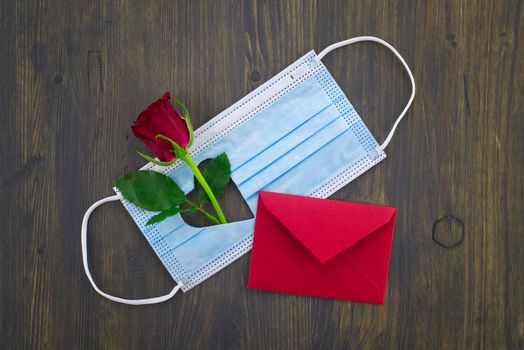 Face mask with cut out heart shape and red rose alongside red envelope on wooden background with free copy space. Social distancing and contact-less greetings