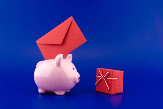 Festive red envelope being inserted into a pink piggy bank next to a present box over blue background with copy space