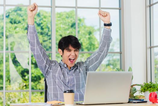 Yes! Happy excited Asian man raising his arm up to celebrate celebrating success. Young businessman using laptop computer at office desk he glad to receive good winner profits from the job