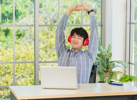 Asian young business man smile listening music in red headphone at home office. Happy businessman sitting on desk and relax wear wireless head phones enjoy listen music and work with laptop computer