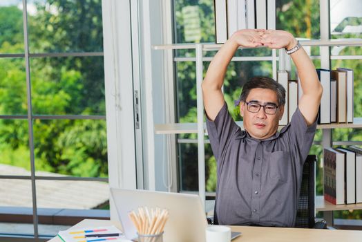 Asian businessman tired overworked he stretch oneself on the desk. senior man with eyeglasses break stretching his arms on table at his working place