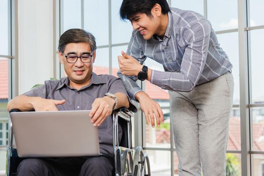 Asian senior disabled businessman in a wheelchair with laptop computer discuss together with team in office. Old father man sitting wheelchair and his son talking video calls conference on laptop