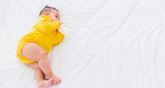 Asian beautiful little baby girl wearing a yellow dress eat milk sleeping feeding lying on the white bed, infant holding a bottle of milk, baby food concept