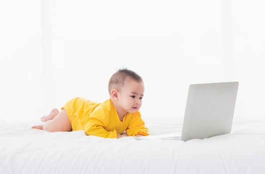 Asian little baby girl wearing a yellow dress typing hands on laptop computer keyboard and looking at monitor on the white bed. Little IT guy