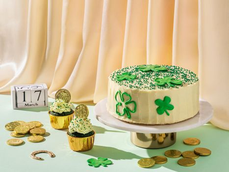 St Patrick's Day food concept. Modern still life with sweet food for Saint Patrick's Day party. Cake decorated shamrocks, green velvet cupckakes, golden coins and horseshoe on golden satin background