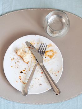 dirty plate after eating on the dining table. Top view of dirty white plate with crosswise cutlery over blue linen tablecloth. Vertical