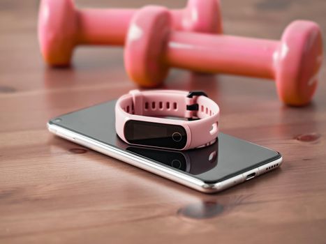 Fitness tracker on smartphone and pink colored dumb-bell on wooden tabletop. Pink female pedometer or wearable device on smartphone with infinity display, copy space