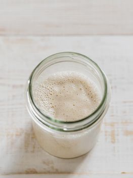 Wheat sourdough starter. Top view of glass jar with sourdough starter on white wooden background. Copy space for text or design. Vertical.