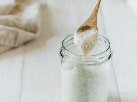 Ready for making bread wheat sourdough starter dripping from wooden spoon. Glass jar with sourdough starter higher hydration on white wooden background. Copy space. Homemade breadmaking concept