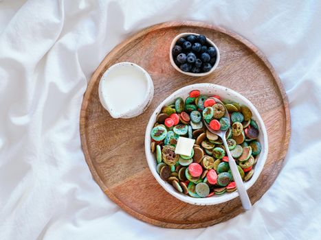 Trendy food - pancake cereal. Bowl of colorful mini cereal pancakes on wooden tray over bed. Tiny pancakes with natural color - green matcha, turquose spirulina, blue pea, red fragon fruit