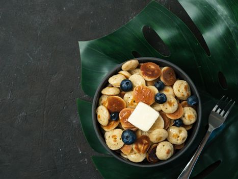 Trendy food - pancakes cereal served butter and blueberries. Heap of mini cereal pancakes in boul on tropical monsterra leaf over dark background. Copy space right for text. Top view or flat lay.