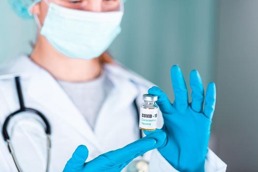 Woman doctor or nurse in uniform and gloves wearing face mask protective in lab holding medicine vial vaccine bottle with COVID-19 Coronovirus vaccine label