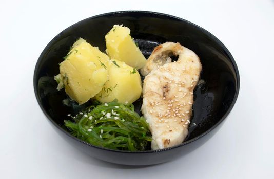 Fried sea fish steak boiled potatoes Chuka seaweed salad on a black plate.