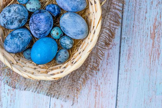 Blue Easter eggs in a rustic, natural style. Eggs in a nest of hay in a wicker basket on burlap on an light wooden background. Copy space.