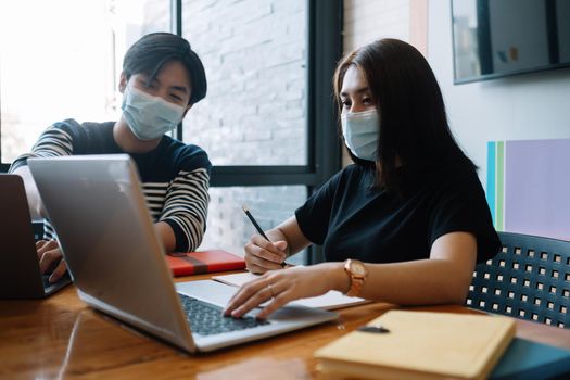 Asian woman and man workers meeting together with laptop for financial and wear protective masks prevent corona virus or covid19 at co working space .Health and teamwork concept