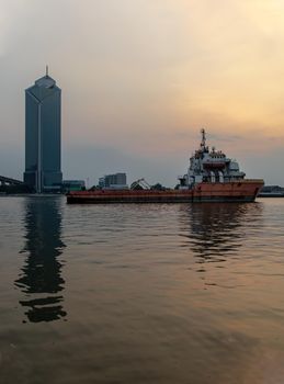 A cargo ship parked in the middle of the Chao Phraya River at evening with Modern building at the background. Selective focus.