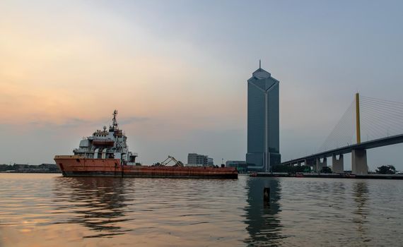 A cargo ship parked in the middle of the Chao Phraya River at evening with Modern building at the background. Selective focus.