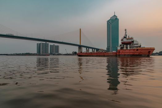 A cargo ship parked in the middle of the Chao Phraya River at evening with Modern building at the background. Selective focus.