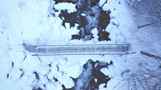 A metal bridge among tall fir trees in the snowy mountains. The view from the drone to the gorge with the river and the trees covered with snow. Clean water, forest. Steep cliffs and rocks. Almarasan