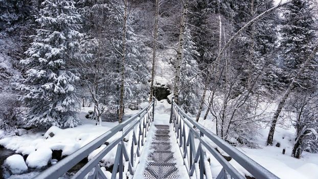 First-person view of a metal bridge in the forest. I walk across a bridge in a snowy gorge, among white rocks and trees. A clear river runs. Mountainous terrain. Snow is falling. Almarasan, Almaty