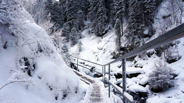 First-person view of a metal bridge in the forest. I walk across a bridge in a snowy gorge, among white rocks and trees. A clear river runs. Mountainous terrain. Snow is falling. Almarasan, Almaty