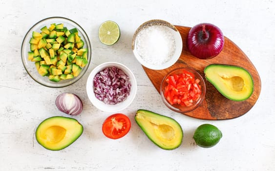 Avocado halves, pieces, limes, tomato and onions, cup with salt - basic guacamole ingredients on white working board, flat lay photo.