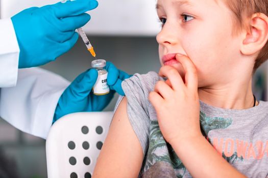 Woman doctor or nurse in uniform and gloves wearing face mask protective in lab, making an injecion to a kid holding vaccine bottle with COVID-19 Coronovirus vaccine label