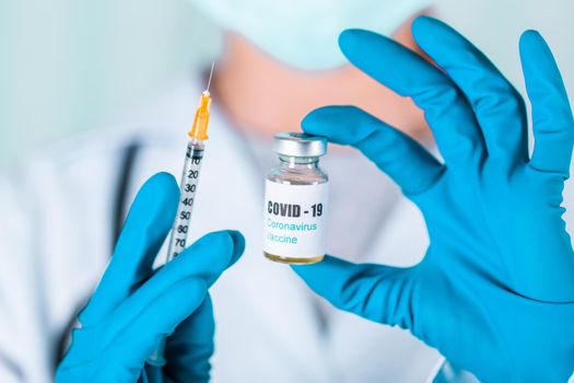 Woman doctor or nurse in uniform and gloves wearing face mask protective in lab, holding medicine vial vaccine bottle with COVID-19 Coronovirus vaccine label
