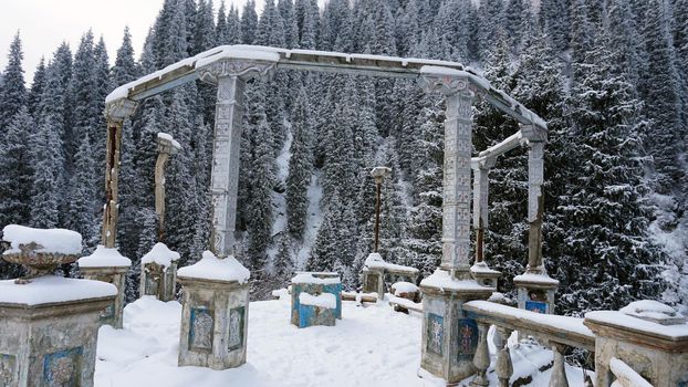 An old abandoned gazebo with views of the snowy mountains and forest. Clouds float beautifully, fir trees are covered with snow. High white hills. The stone pillars of the building are destroyed.